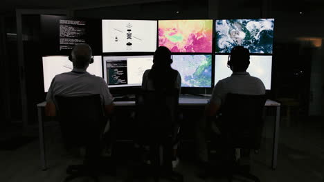 three people working at a computer desk in an office, monitoring data on multiple screens.