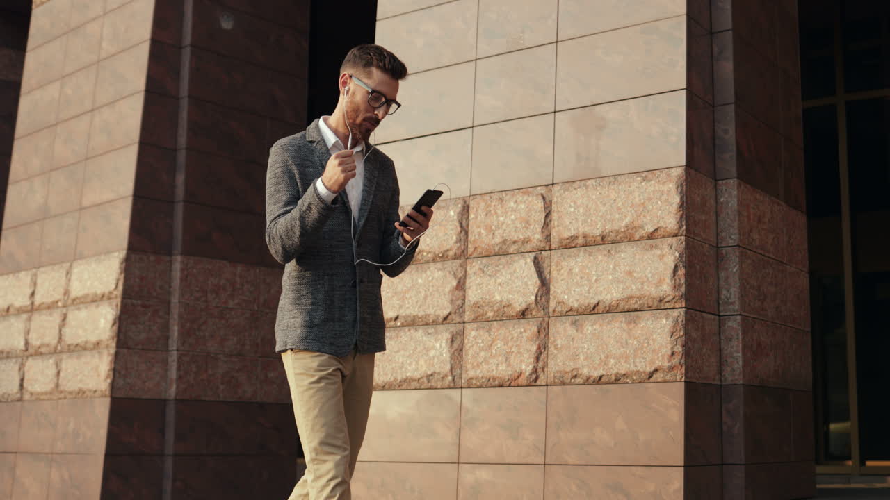 hombre elegante escuchando música en el teléfono inteligente a través de auriculares mientras camina y baila alegremente en la calle 1