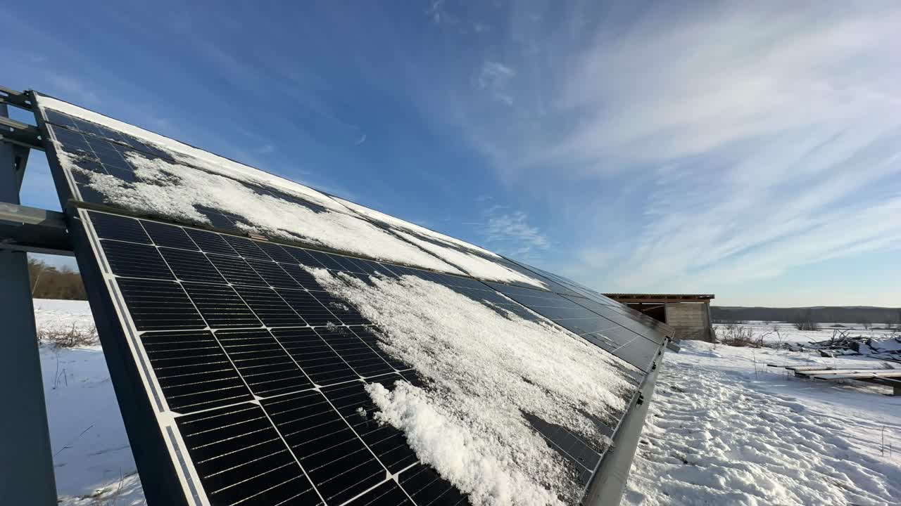 Zimní časosběr fotovoltaických panelů částečně pokrytých sněhem. Mraky se ženou od západu na východ, zatímco slunce vychází a postupně rozpouští sníh, přičemž odhaluje tmavé skleněné řady pod ním.