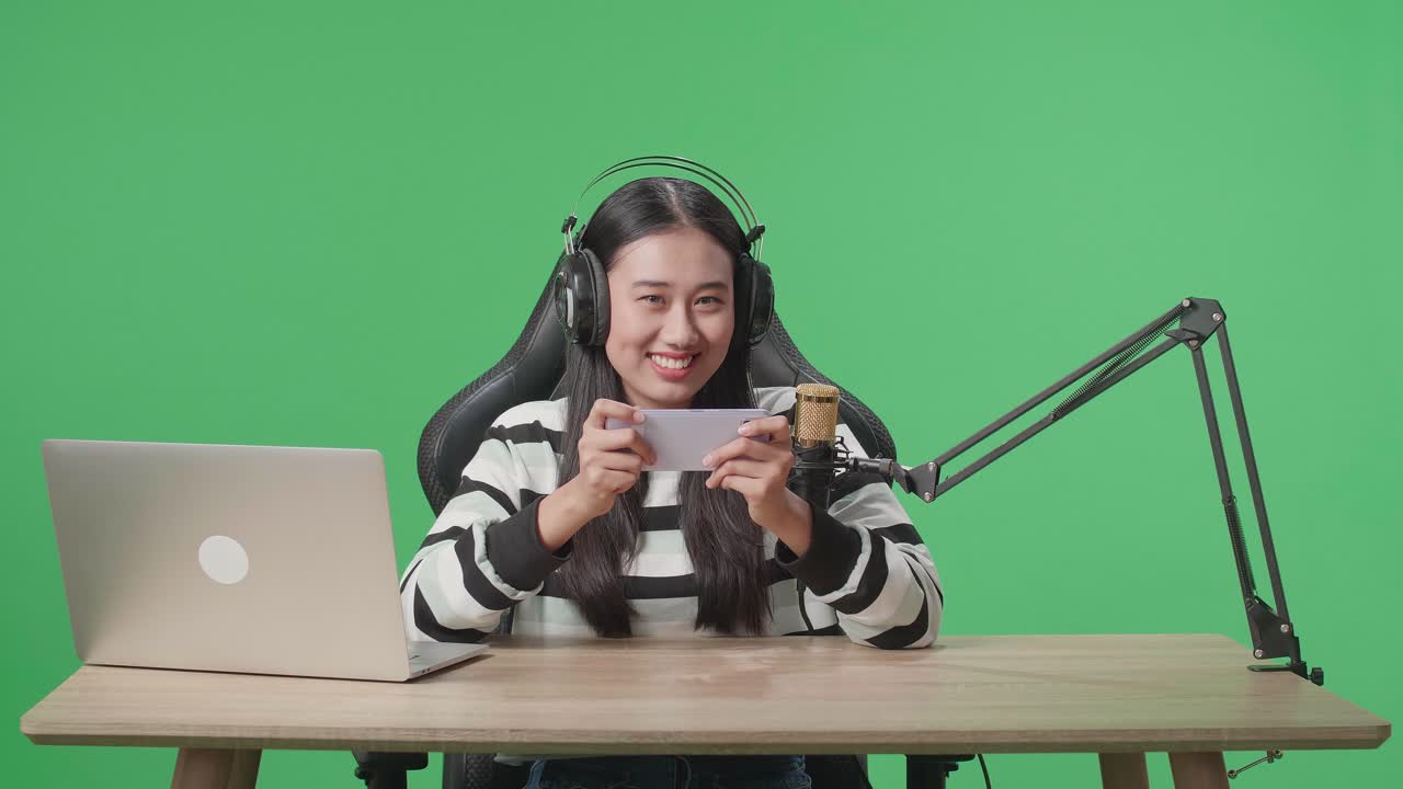 Una donna asiatica gamer sorridente con cuffie e computer, che guarda la telecamera mentre gioca al telefono cellulare su sfondo green screen