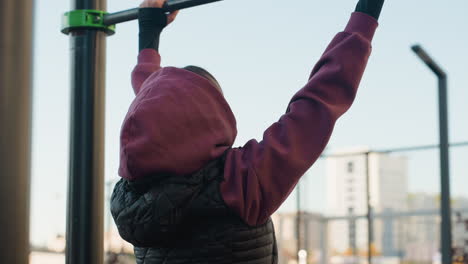 side view youthful woman in burgundy hoodie and black vest places hands on horizontal bar for balance on rubber flooring in urban park outdoor gym with yellow building backdrop conveying form