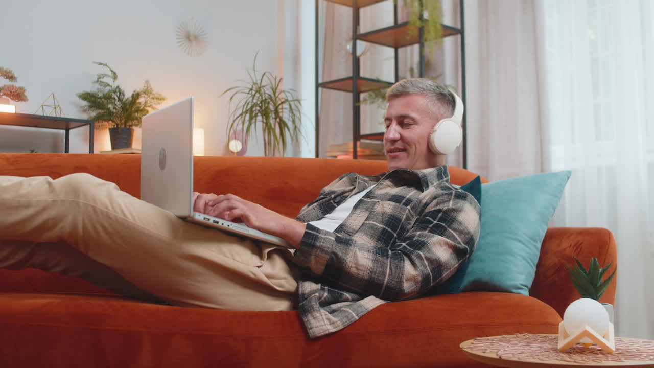 Happy man in wireless headphones lying on home couch listening music working on laptop relaxing