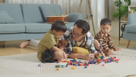 cuerpo lleno de padre y hijos asiáticos jugando el conjunto de construcción ladrillo de juguete de plástico colorido en casa. los niños ensamblan bloques de construcción de plástico sentados en una alfombra, un padre acostado en el suelo hablando con sus hijos