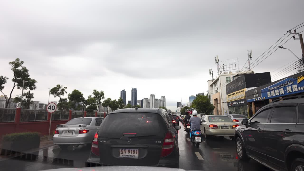 Motorcycles weave through congested cars on wet Bangkok street under overcast sky, wide dashcam view