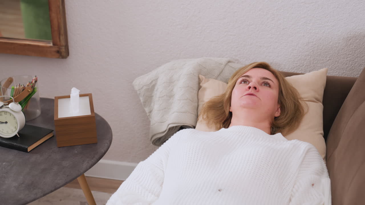 Conversion partner lies faced up on comfortable brown sofa gazing upward while having open conversation with empathetic listener, pillows behind head, nearby table holds clock tissue box pencils