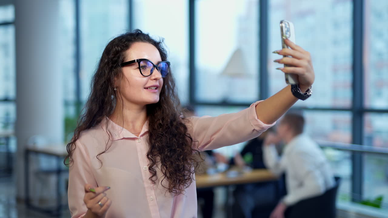 Charming lady with long dark curly hair and glasses takes video on her phone. Office team working at backdrop in blur.