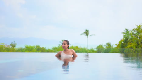 Front-view-of-Smiling-happy-Asian-lady-in-an-infinity-outdoor-swimming-pool-She-is-leaning-her-arms-on-the-edge-of-the-pool-in-Hawai