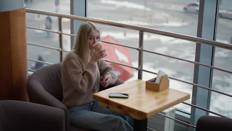 elegant lady sipping coffee from cup while looking out through window, phone resting on wooden table in modern mall cafe with urban setting in background, soft natural lighting and serene mood