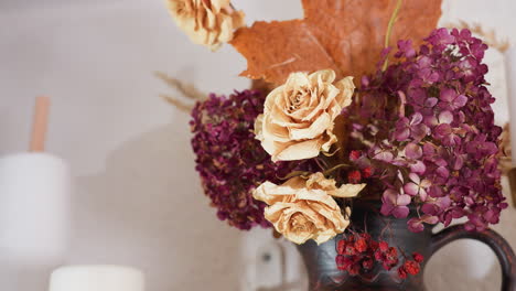 close up of dried flower bouquet with beige roses, deep purple hydrangeas, red berries, and orange maple leaf in dark ceramic vase, set beside household decor and paper towel roll