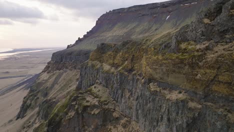 Aerial-landscape-view-of-mountain-rock-formations-and-steep-cliffs,-with-melting-snow,-on-a-moody-evening-in-Iceland