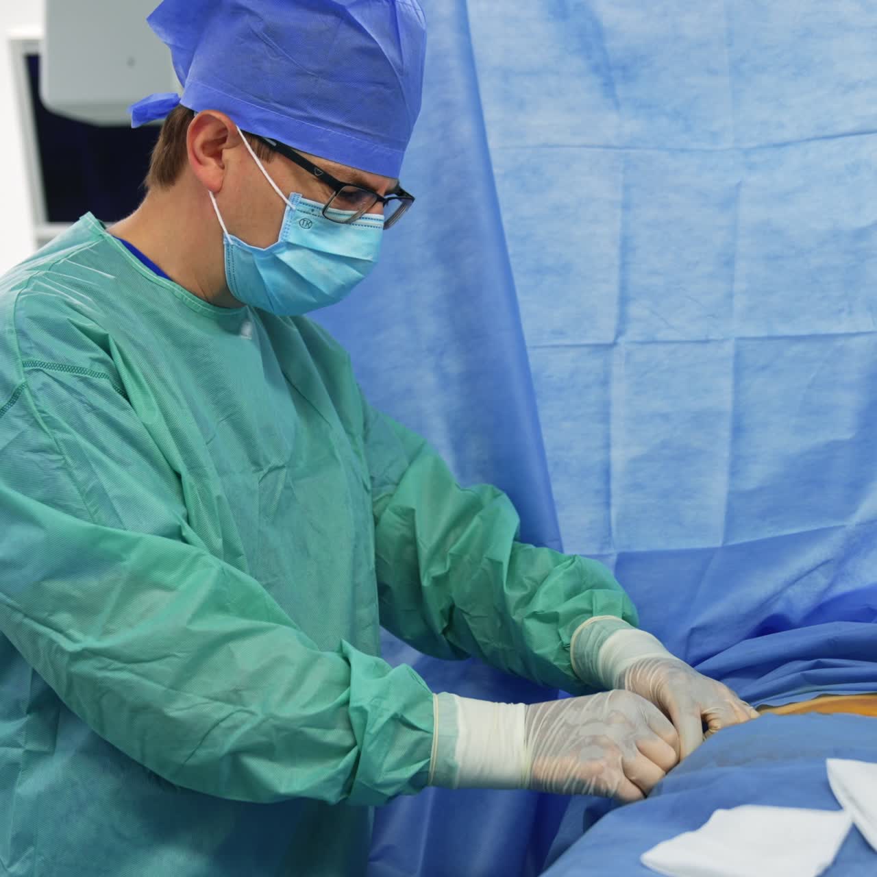 Doctor in blue robe, hat and mask stands at the operational table. Surgeon making cut with a scalpel in the patient's body