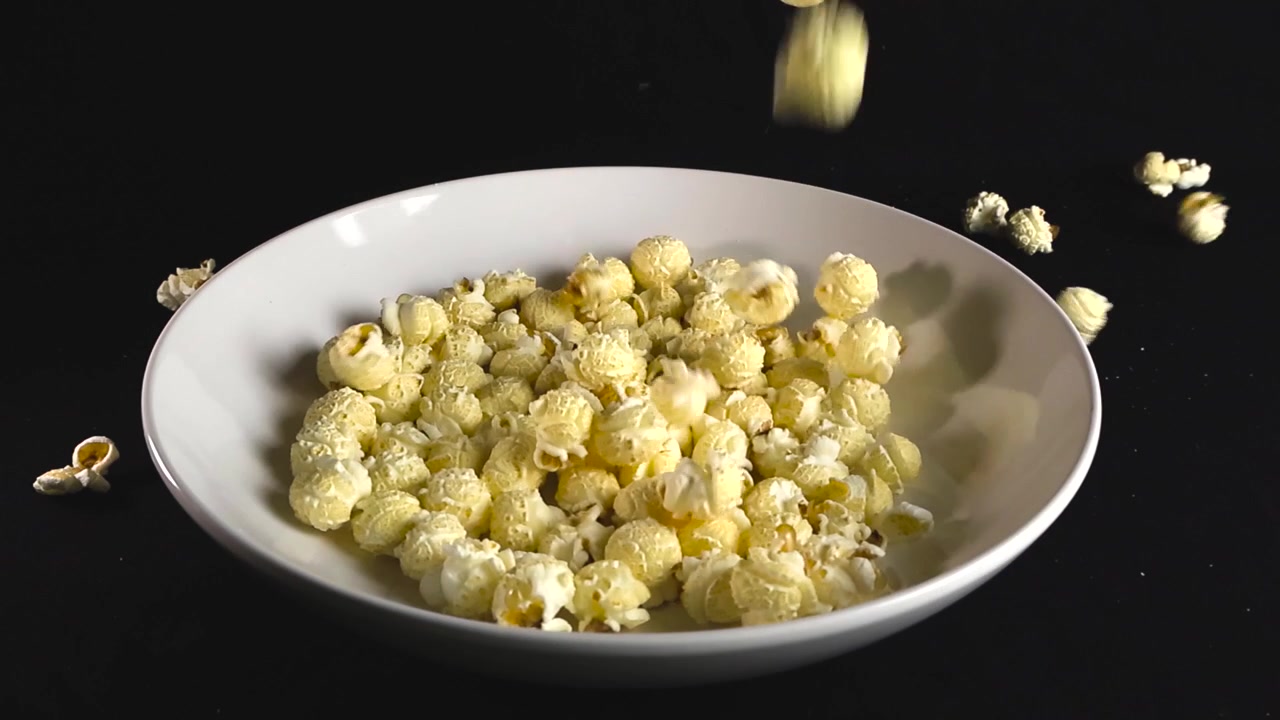 Krydret, luftigt popcorn, der falder ned i en hvid keramikskål mod en ensfarvet sort baggrund – et perfekt visuelt udtryk for snacks, film, fastfood og lækre godbidder.