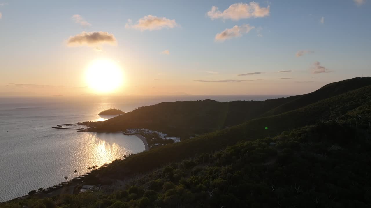 Aproximação lenta de drone ao pôr do sol sobre a crista da Ilha Hayman, revelando a baía abrigada e o resort abaixo com reflexos dourados, Whitsundays, Queensland, Austrália