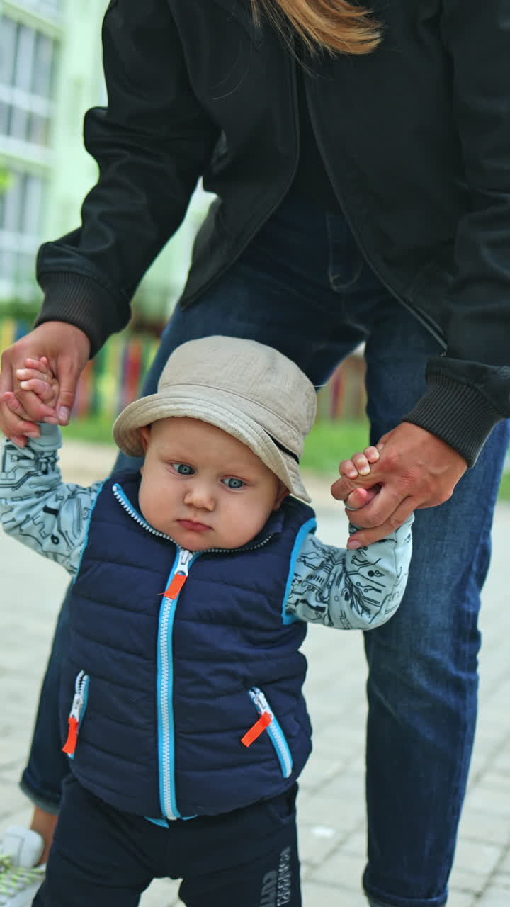 A baby boy learns to walk holding his mother's hand on the street. Baby's first steps. Development of the baby. Clothes for babies. Vertical video