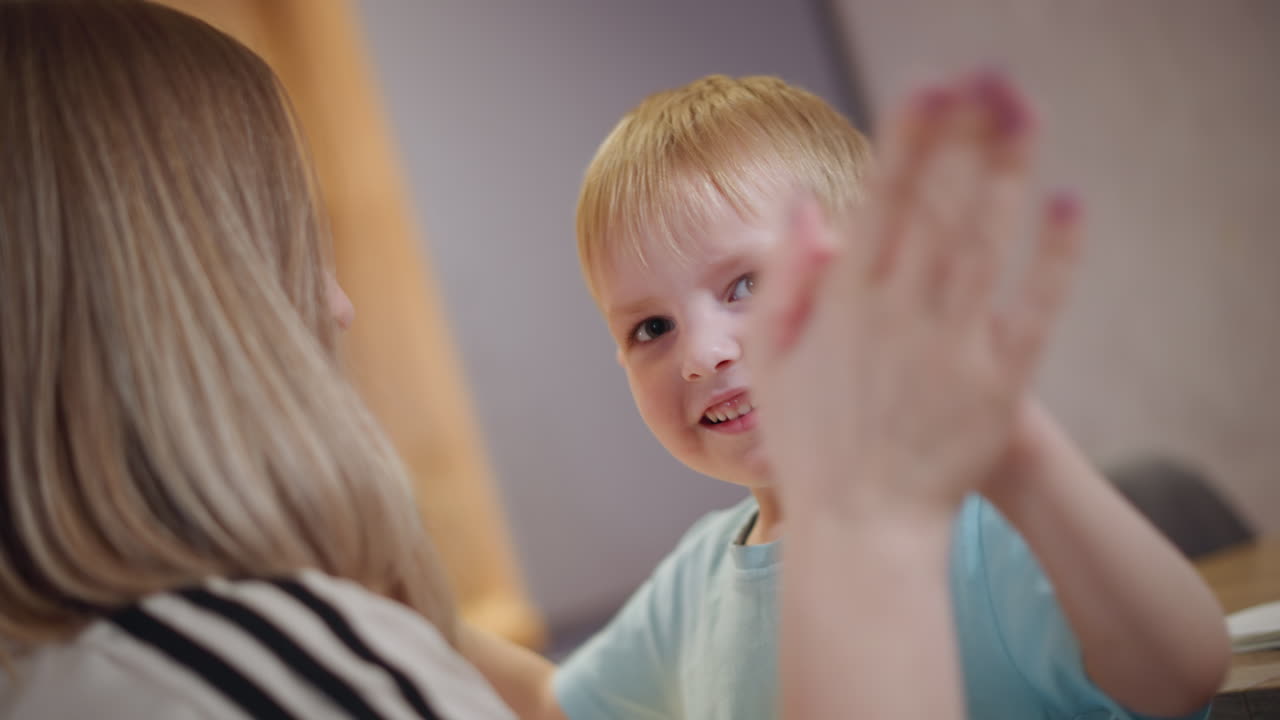 Smiling young boy in blue shirt looks happily toward camera while sitting near mother at home table, showing joy, innocence, warmth, trust, and family bond in casual natural moment filled with love
