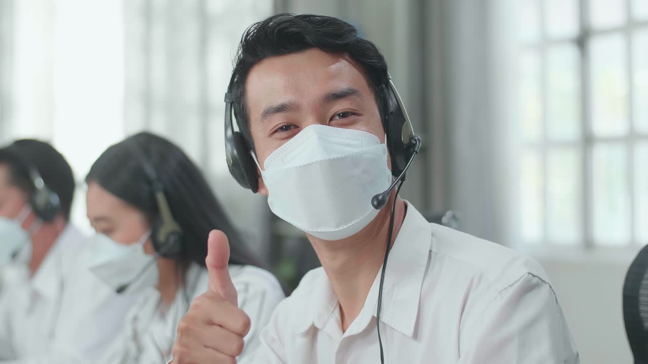 Close Up Of A Man Of Three Asian Call Center Agents Wearing Headset And Mask Looking At Camera And Thumbs Up While Two Of His Colleagues Are Speaking With Customer At The Office
