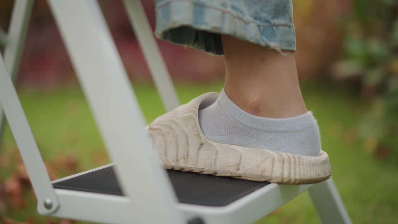Nærbillede af fod, der træder på stigetrin, med afslappet hjemmesko, sok og denimjeans. Metalfolde-stige, græsklædt baghave med efterårsblade. Forsigtig opstigning af husejer, fokuseret balance, huslig pligt, slowmotion.