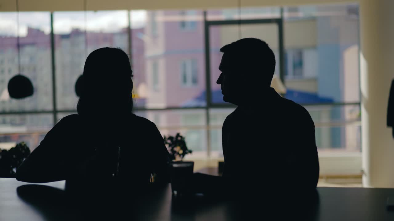 silhouettes of man and woman are talking at a table in the cafe and drinking drinks on the background of the window. Blurred background