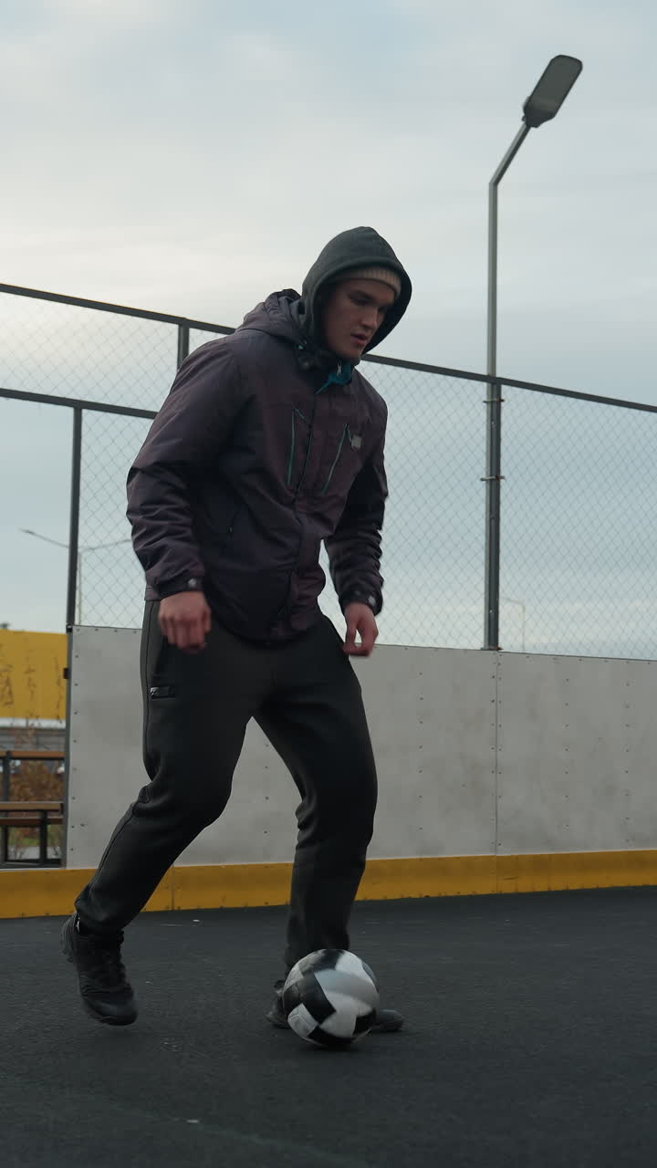 Young athlete skillfully training with soccer ball on sport arena during a practice session, background includes urban buildings, fencing, and goalpost