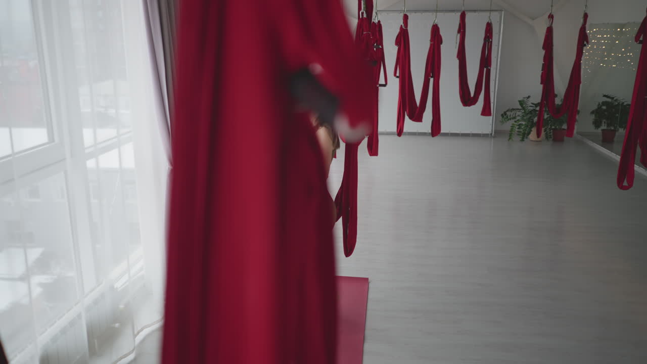Well lit high angle view of wellness guide sitting on red aerial silk hammock in modern yoga studio with glass windows and curtains, holding hammock firmly conveying balance and strength