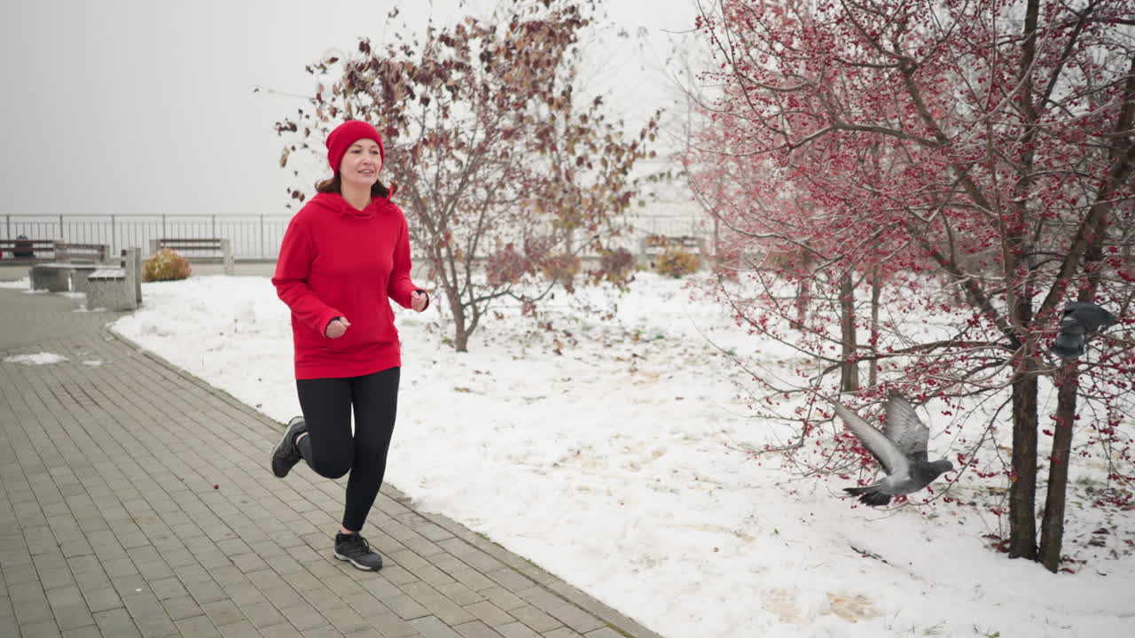 mujer corriendo por un camino pavimentado cubierto de nieve con una cálida sonrisa durante el invierno rodeado de paisajes serenos del parque, árboles estériles y bancos, una paloma vuela cerca mientras se acerca exuding energía