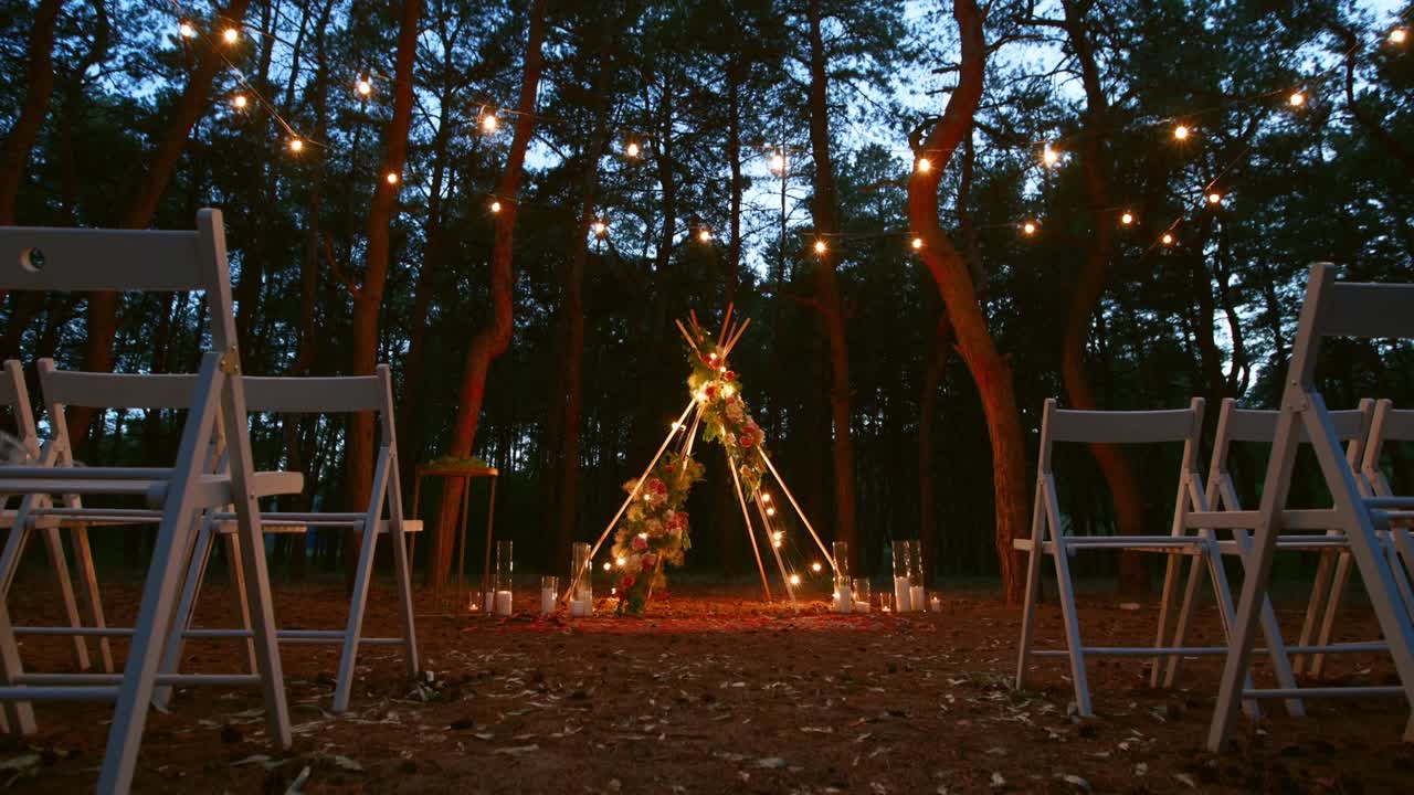luces de cadena festivas iluminación en el arco de boho tipi decoración en el lugar de la ceremonia de boda al aire libre en el bosque de pinos por la noche. luces de cuerda vintage guirnaldas de bombillas que brillan sobre las sillas en la boda rural de verano