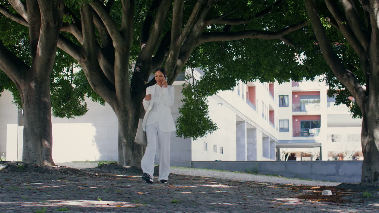 богатая леди, говорящая на смартфоне, гуляет по городской аллее в белом костюме. женщина звонит.