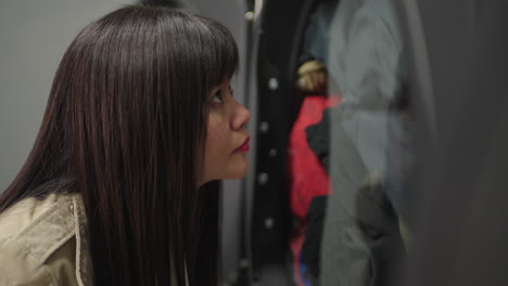 A-contemporary-Asian-woman-with-bangs-leans-against-the-laundry-door,-her-hand-resting-on-her-forehead,-as-she-waits-patiently-for-her-clothes-to-finish-washing