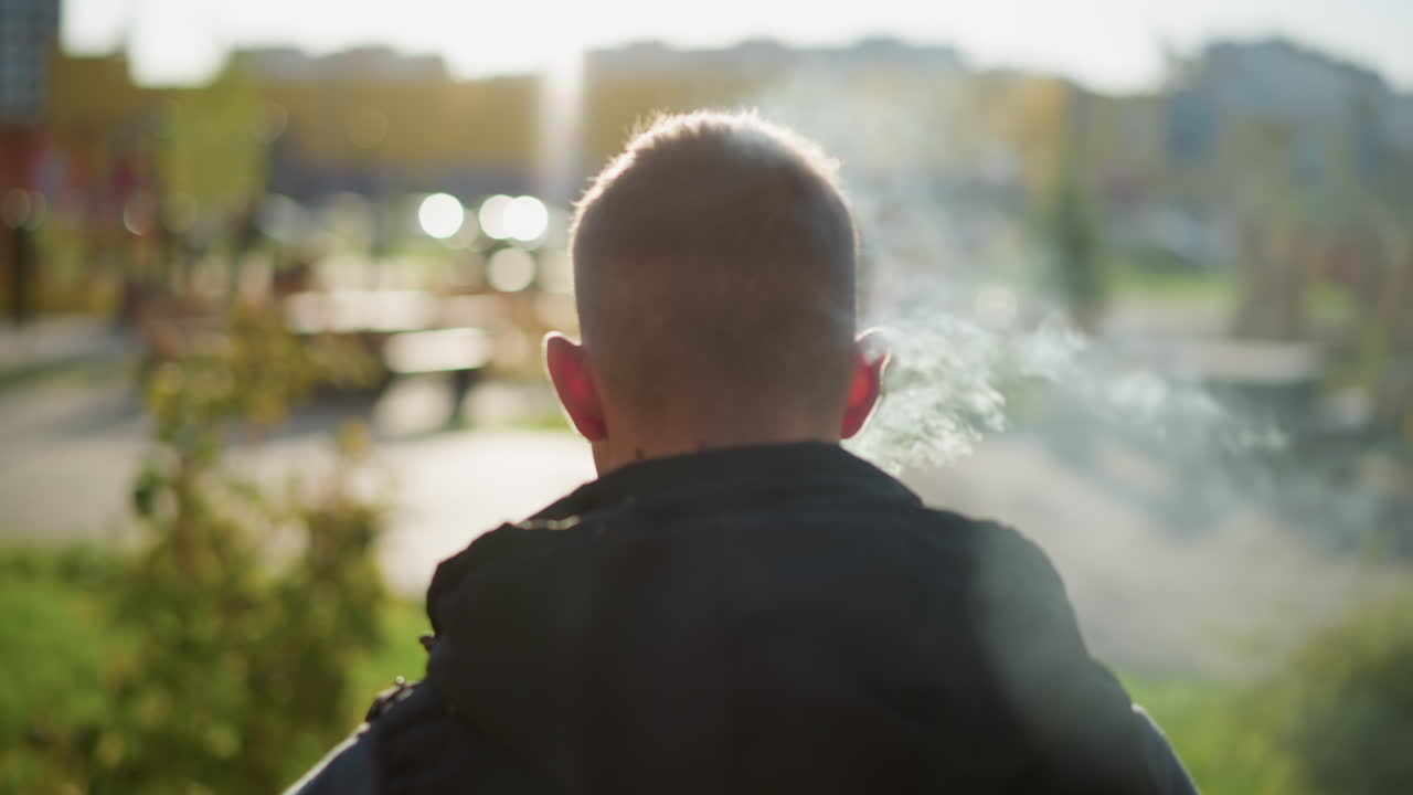 back view young man stands in sunny park exhaling dense white shisha smoke into air while facing vibrant urban background with blurred buildings and trees during bright afternoon light