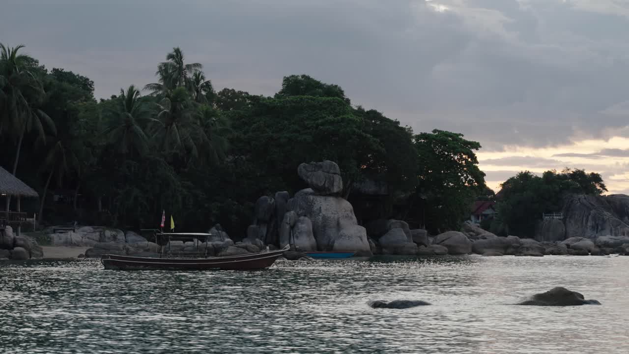 A wooden longtail boat floats near rocky formations and palm trees on Ko Tao at dusk