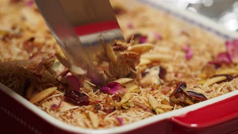 serving a traditional vermicelli dessert, possibly kunafa, garnished with nuts and rose petals from a baking dish