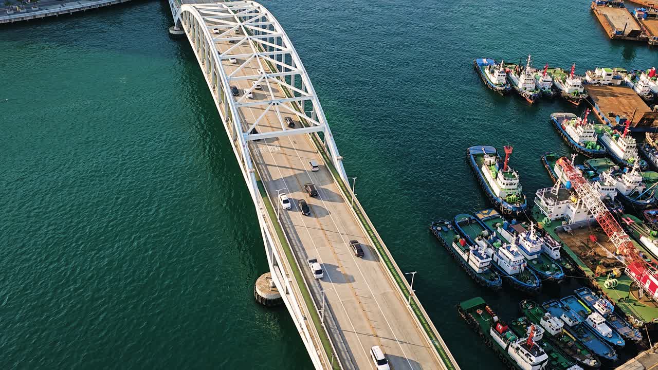 Drone view of Busan Bridge in South Korea showing vehicles crossing above blue waters with moored ships alongside the harbor