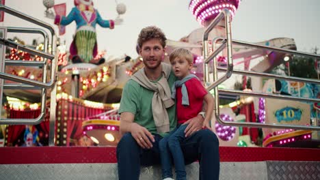 un padre con cabello rizado con paja en una camiseta verde se sienta cerca de su pequeño hijo rubio en una camisa roja que se sienta en su pierna y mira las atracciones en un parque de atracciones con luces brillantes