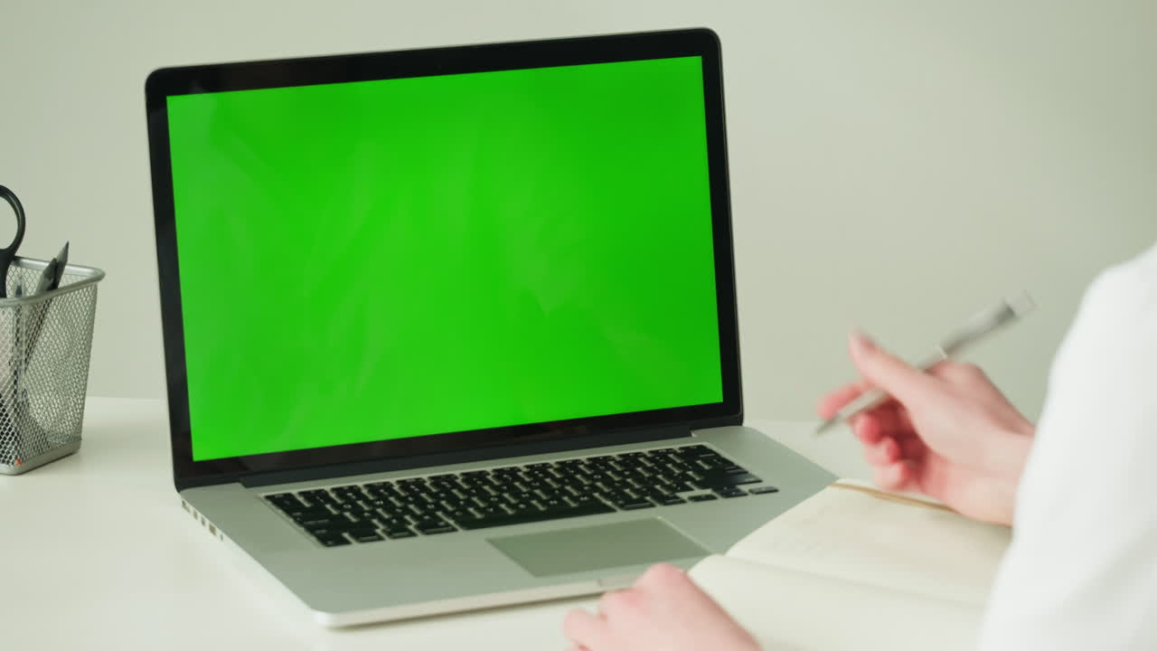 Person working on a laptop with a green screen, taking notes in a notebook.