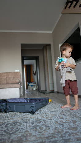lovely toddler walks around the suitcase holding a pile of his clothes. happy kid tosses his things up smiling. low angle view. vertical video.