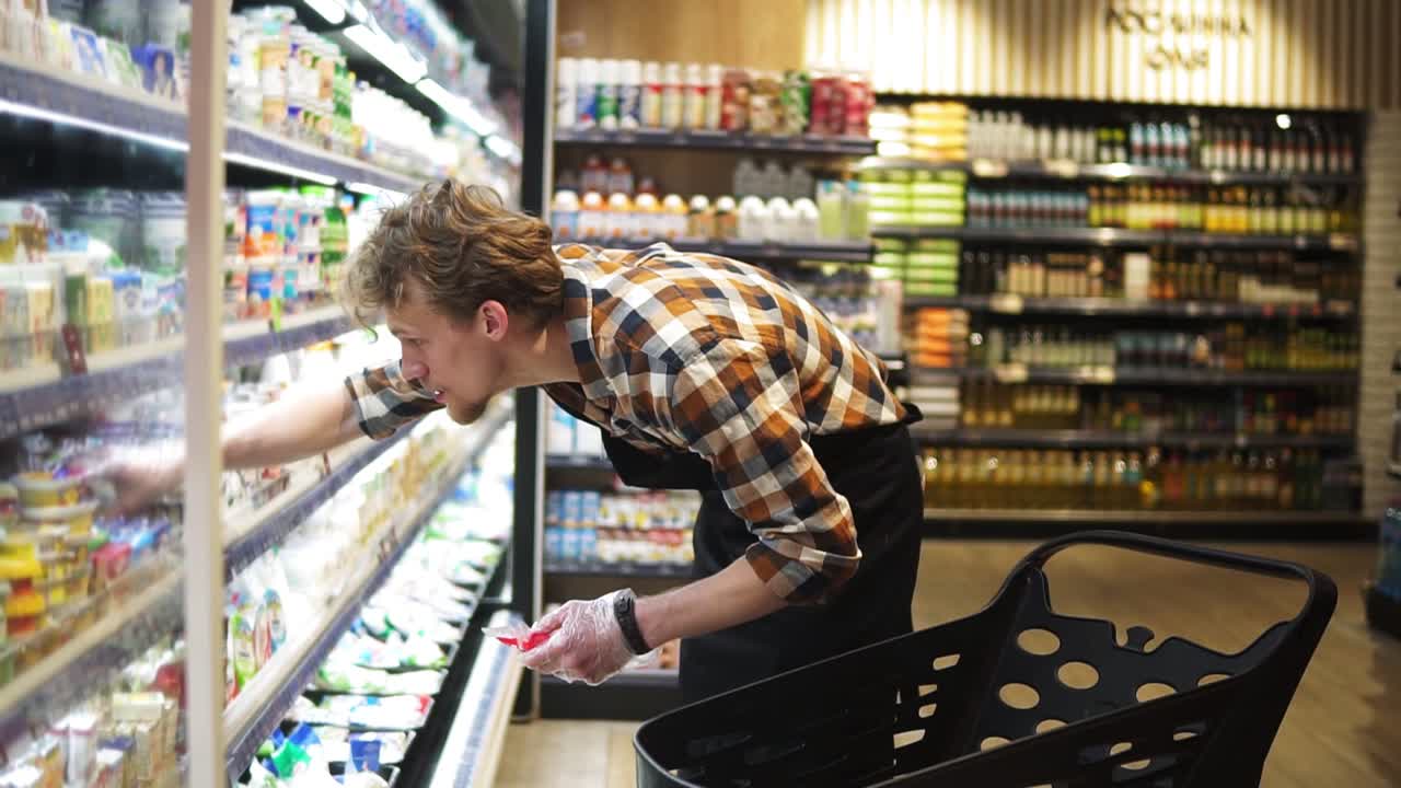 Salesman is putting cheese packs from the cart on shelves in milk products department in food store, bearded guy is wearing apron. Selling products, profession and trade concept