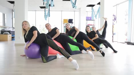 A group of women in sportswear are leisurely stretching their legs, knees, and thighs with pilates balls in a gym building