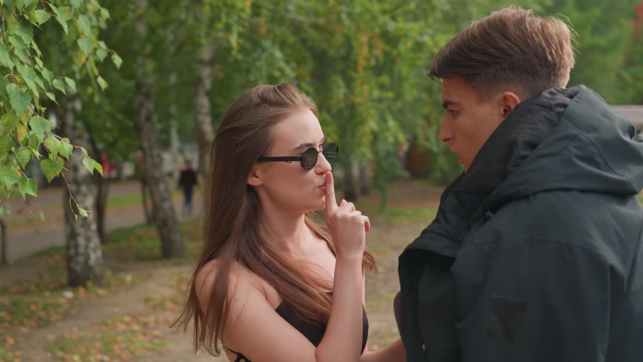 Mladá žena pomáhá třesoucímu se muži s bundou, scéna venkovního parku s lavičkami a stromy, sluneční brýle a letní šaty kontrastují s nadměrným kabátem, něžné ruce upravují límec, hravá útěcha