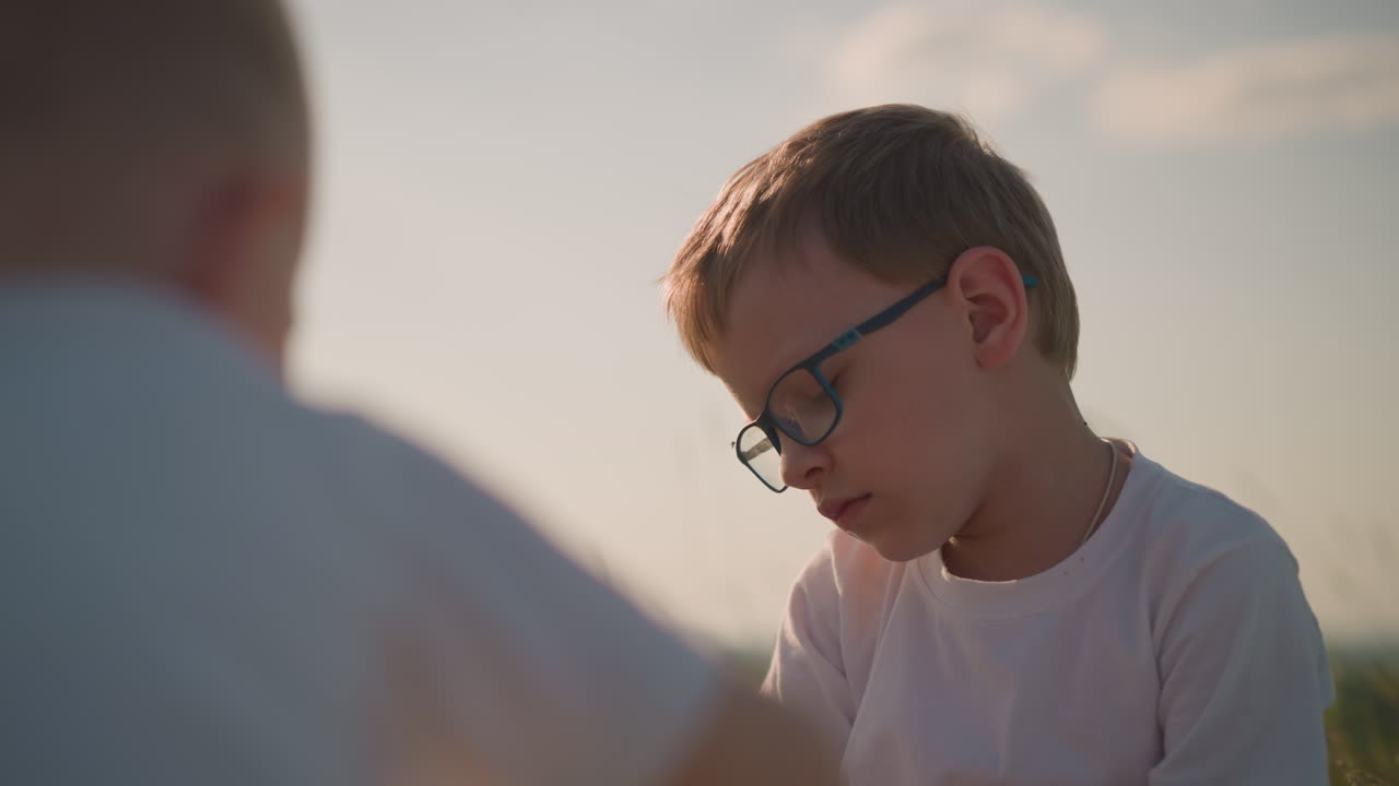 A close-up of a boy wearing glasses and a white shirt, with a thoughtful and unhappy expression on his face. His brother is blurred in the background, adding depth to the scene