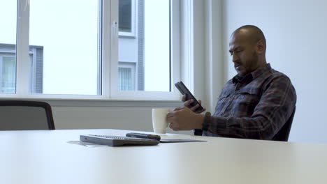 Lone-Bald-Bearded-Indian-Male-Taking-Seat-In-Meeting-Room-Putting-Mug-On-Table-And-Checking-His-Phone