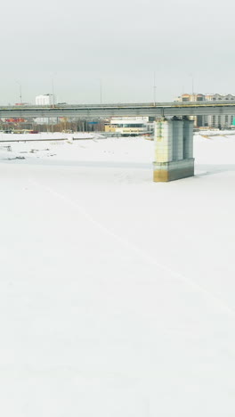 person silhouette walks along white snowy path on frozen river against bridge and modern buildings upper view