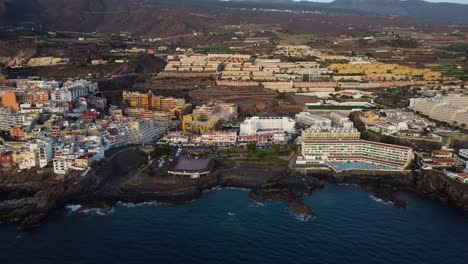 beautiful drone shot seashore seaside of spain tenerife island volcano in the background and mountains water sea blue coast hotel pool buildings los gigantes