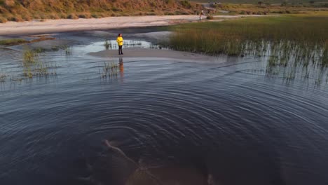 Aerial-orbit-shot-of-Young-man-standing-on-lagoon-shore-island-at-sunset-and-enjoying-lake-view---Laguna-Negra,Uruguay