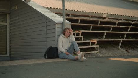 young lady sitting on ground near stadium walkway, resting head on hand, leaning on pole, lost in thought, black bag beside her, deep in quiet reflection