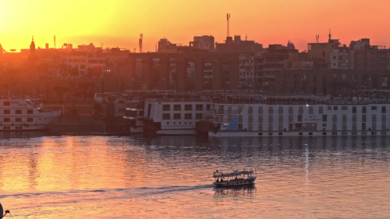 Una serena vista all'ora d'oro di una piccola barca passeggeri che scivola sulle calme acque del fiume Nilo, con grandi navi da crociera ormeggiate lungo la riva illuminata di Luxor.