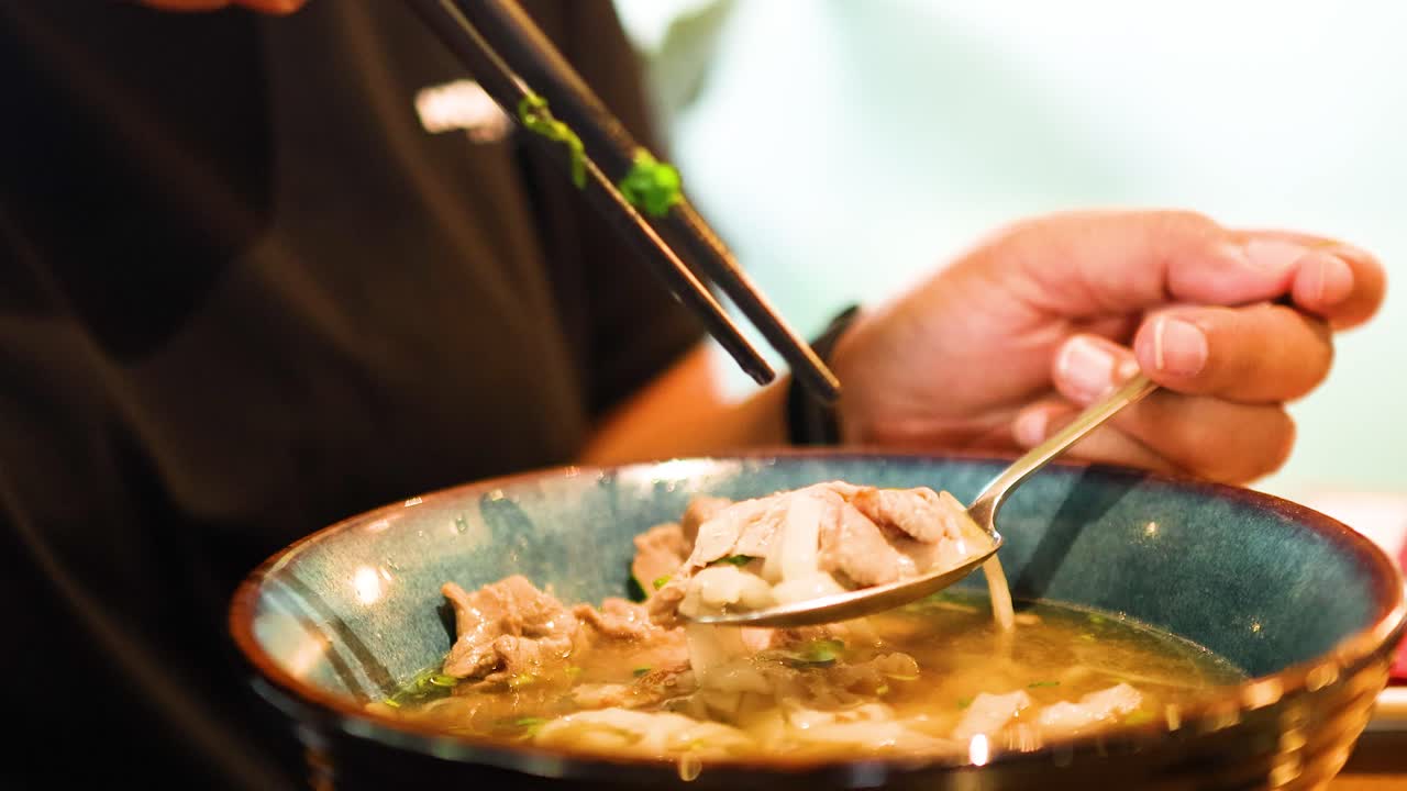 A person savors a bowl of Vietnamese pho using chopsticks and a spoon in a warmly lit Bangkok restaurant