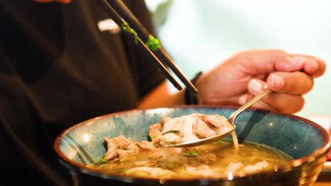 a person savors a bowl of vietnamese pho using chopsticks and a spoon in a warmly lit bangkok restaurant