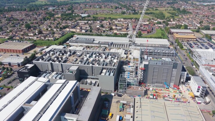 Aerial view captures a massive data center complex undergoing active construction on a clear, sunny day. Multiple buildings are taking shape with cranes and equipment present.