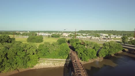 Flight-above-steel-truss-railroad-bridge-spanning-across-brown-river-toward-horizon,-aerial-overhead-approach
