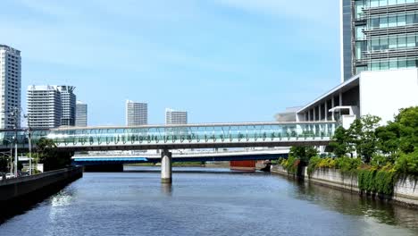 Modernes-Stadtbild-Mit-Einer-Eleganten-Fußgängerbrücke-über-Einen-Ruhigen-Fluss-An-Einem-Sonnigen-Tag